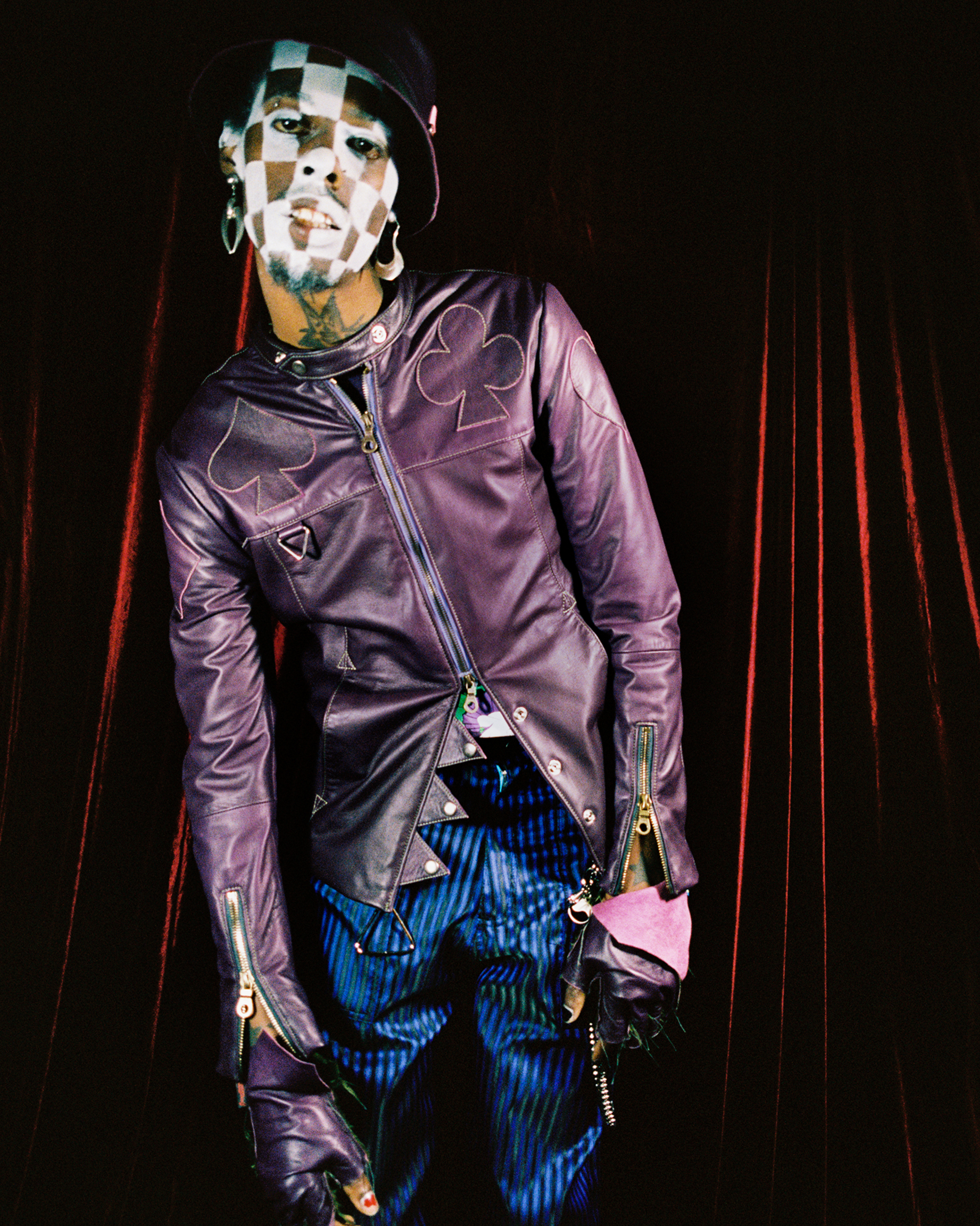 Deep-skined model with white checkered makeup leaning towards the camera in a pruple jacket and blue pants against a red velvet background